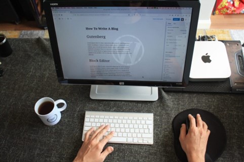 Computerskærm, åben på en WordPress side, med hænder på mus og tastatur og en kaffekop ved siden af